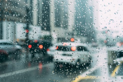 Raindrops on a window blur a view of a busy city street with several cars and red tail lights visible. The scene depicts an overcast day with a sense of motion and wet conditions.