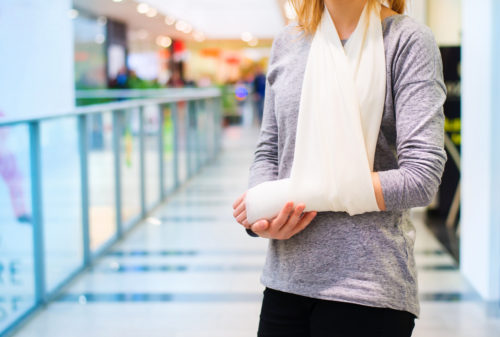 A person in a gray long-sleeve shirt stands in a mall, holding their arm in a white sling. The background shows a blurred shopping area with glass railings.