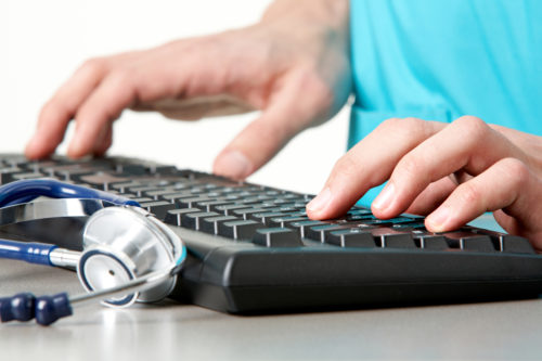 Close-up of hands typing on a keyboard with a stethoscope lying nearby on a desk, suggesting a healthcare or medical context.