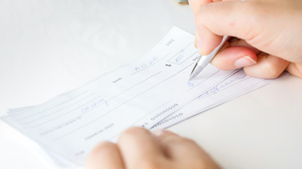 Close-up of a person's hand holding a pen and writing on a stack of checks or forms, possibly documenting lost future wages, with handwritten text visible on the top sheet. The background is white and out of focus.