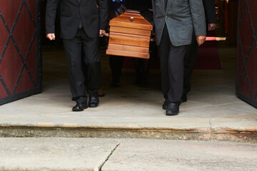 Two people in formal attire carry a wooden coffin out of a building, possibly during a funeral service, after a fatal car accident, as they walk through large open doors.