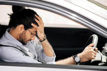 A man with glasses sits in a company car, holding the steering wheel with one hand and resting his head in his other hand, appearing stressed or frustrated, possibly after an accident.