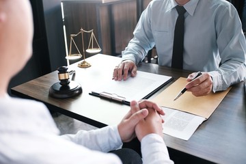 Two people sit across a desk, one in a suit holding a pen with documents, a gavel, and scales of justice on the table, indicating a lawyer consultation or legal meeting.