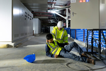 A worker in a reflective vest and helmet checks on a colleague who has suffered a construction injury at an industrial site, with a blue hard hat lying nearby.