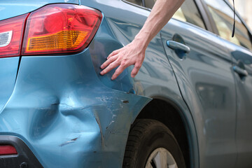 A person’s hand reaches toward the dented and scratched rear fender of a blue car, assessing visible property damage near the wheel and tail light—potential grounds for compensation.