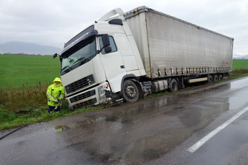A large white truck, possibly a victim of causes of truck accidents, is stuck in a grassy ditch beside a wet road. A person in a yellow rain jacket stands nearby as rain falls and skies remain overcast.
