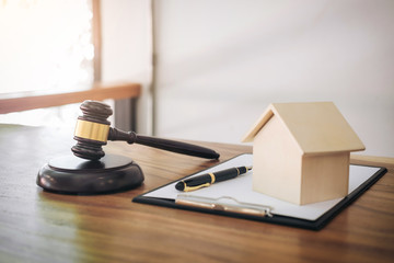 A wooden gavel, a model house, a pen, and a clipboard with paper are arranged on a wooden desk, suggesting a legal or real estate setting involving landlords or building code violations.