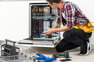 A man wearing a tool belt kneels in front of an open dishwasher, inspecting the inside. Tools, gloves, and a removed rack are on the floor nearby, as he checks if the manufacturer is liable for any faulty appliances.