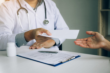 A doctor in a white coat hands a prescription or medical advice to a patient across a desk with a clipboard, pen, and medicine bottle on it.