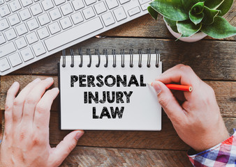 A person writes "PERSONAL INJURY LAW" on a spiral notebook, placed on a wooden desk beside a keyboard and a small potted plant, considering how to file a lawsuit and get compensated for damages under PIP coverage.