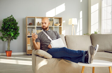 A man with a beard sits on a sofa, his arm in a sling from an accident, looking at his smartphone. Relaxed with legs stretched out, he’s likely searching how to recover lost wages in his bright, modern living room.