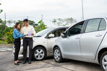 A man and woman stand between two silver cars that appear to have been in a minor accident. They are looking at documents, likely involved in an information exchange after accident, with both cars parked close together on a street.