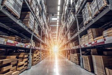 A brightly lit warehouse aisle with tall shelving stacked with large boxes and packages, creating a symmetrical pattern. Safety is key to prevent injured workers from falling merchandise as sunlight streams in at the vanishing point.