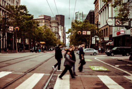 People walk across a busy city street at a crosswalk, surrounded by tall buildings, trees, cars, and storefronts under a cloudy sky. The scene appears urban and lively.