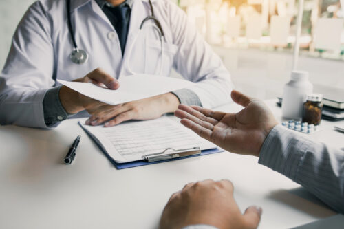 A doctor in a white coat hands a document to a patient across a desk. The patient gestures with an open hand, and a clipboard with papers, a pen, and medicine bottles are on the table.