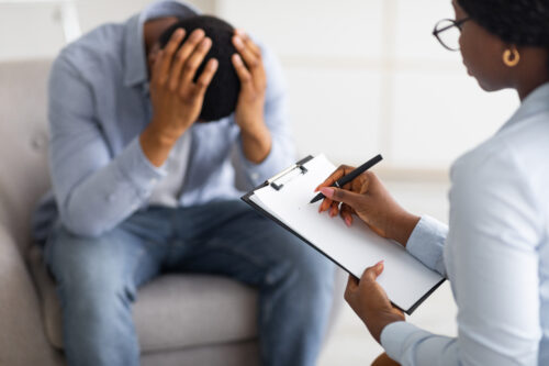 A person sits on a couch with their head in their hands, appearing distressed, while another person holds a clipboard and pen, seemingly taking notes or conducting a counseling session.