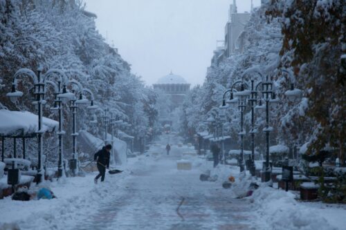 A snow-covered city street lined with trees and lamp posts, with a person shoveling snow in the foreground and another walking in the distance; a domed building is visible at the end of the street.