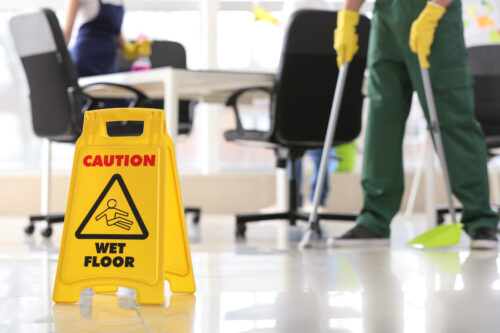 A yellow "Caution Wet Floor" sign stands on shiny tile. In the background, two people in cleaning uniforms and gloves mop the floor near office desks and chairs.