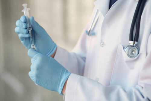 A healthcare professional in a white coat and blue gloves prepares a syringe, drawing liquid from a vial. A stethoscope hangs around their neck.