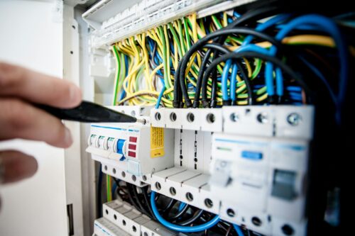 A hand using a tool to adjust switches in an electrical panel filled with colorful wires and circuit breakers.