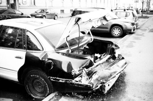 A heavily damaged car with its trunk and rear end smashed is parked on a city street next to other vehicles. The car’s back is crumpled and exposed, indicating it was in a severe collision.