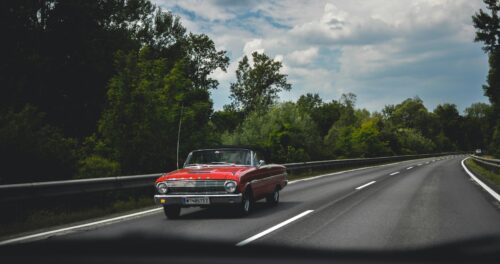 A red vintage convertible car with its top down drives on a curving, empty highway surrounded by trees and greenery under a partly cloudy sky.