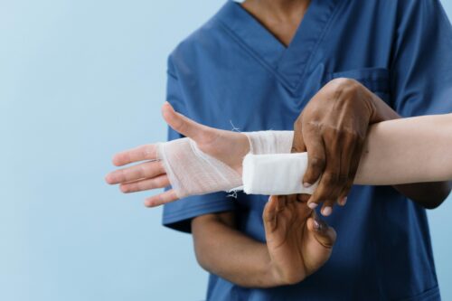 A healthcare worker in blue scrubs wraps a bandage around a patient's injured wrist and hand against a light blue background.