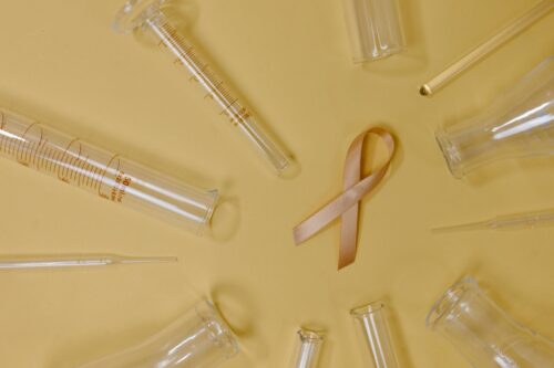 Clear laboratory glassware and pipettes arranged in a circle around a gold awareness ribbon on a yellow background, symbolizing scientific research and support for a cause.