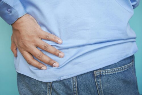 A person wearing a blue shirt and jeans places their hand on their lower back, suggesting discomfort or pain in that area. The background is a solid light blue color.